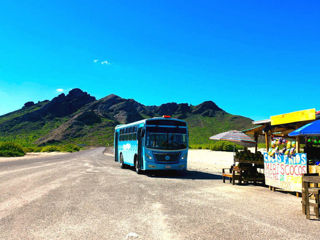 A local blue bus parked in front of mountains near a coconut stand - taking public transport is the best way to travel on a shoestring!