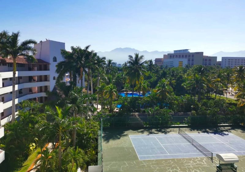 A tennis field surrounded by green palm trees, condos and blue sky - one of our free accommodations in Mexico!