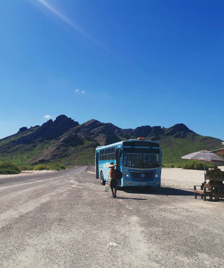 A local bus parked in front of a mountain at a beach - taking public transport is one of the best ways to travel cheap!