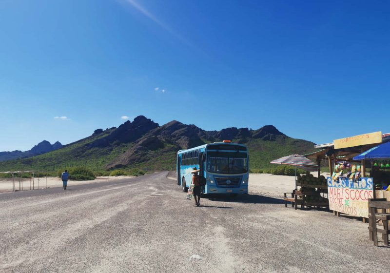 A local bus parked in front of a mountain at a beach - taking public transport is one of the best ways to travel cheap!