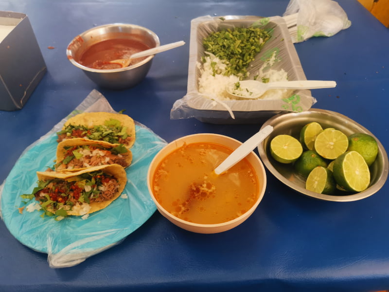 Plate of tacos next to a bowl of soup, a plate of onion, cilantro and a bowl of limes