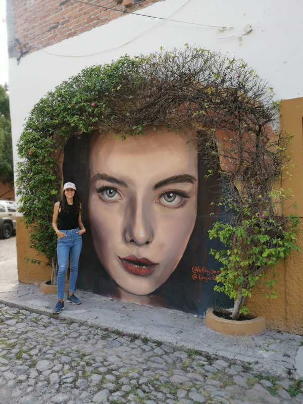 Katharina standing next to an unusual street art of a womans face surrounded by plants as hair in Guadalupe 