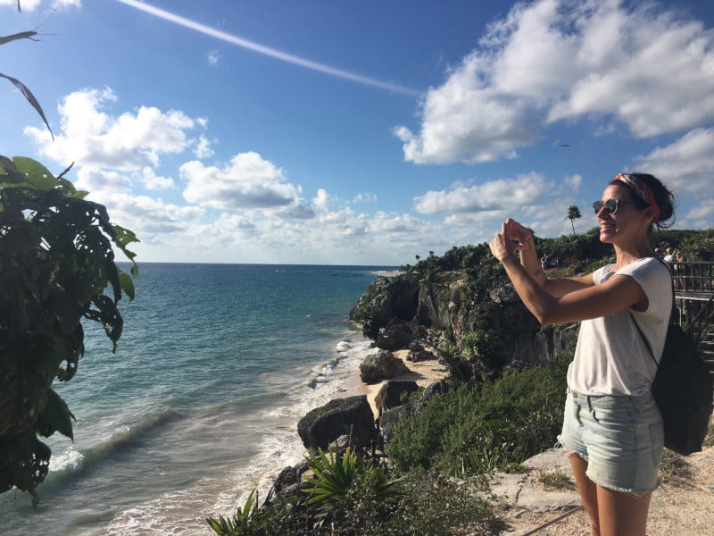 Katharina standing next to the ocean at Tulum ruins forming her hands to a heart