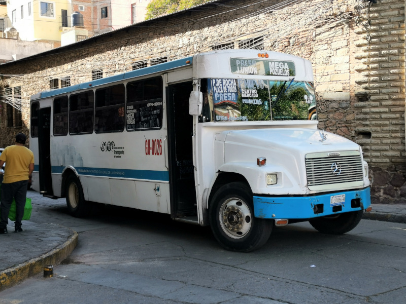 A white and blue Guanajuato bus standing at a bus stop with destinations written on its windshield