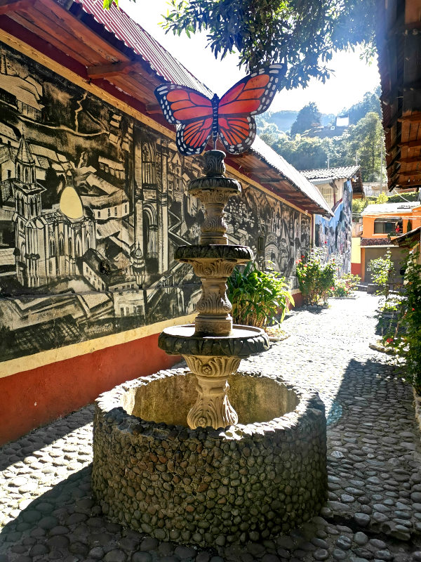 A fountain in the middle of an alleyway with a butterfly mural on the wall in Agangueo Mexico