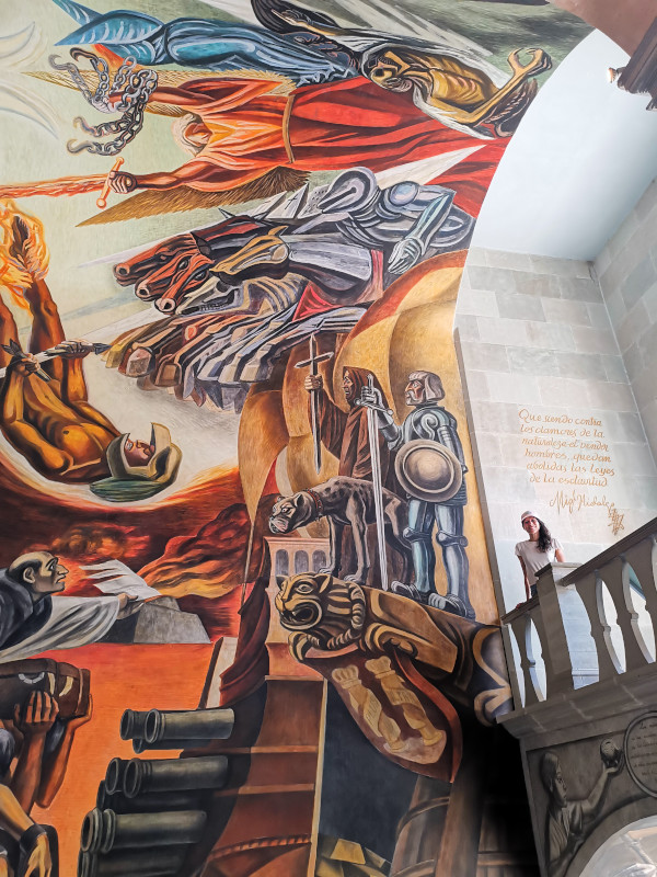 Katharina standing on a balcony overlooking a large colorful mural inside the regional museum of Guanajuato