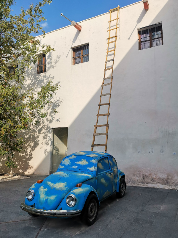 An art display with a ladder coming out of the top of a VW car painting in blue with white clouds
