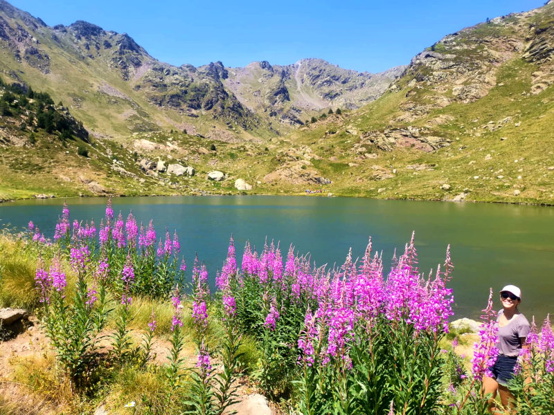 Katharina standing next to a lake with some pink flowers in the foreground on the Estanys de Tristiana hike