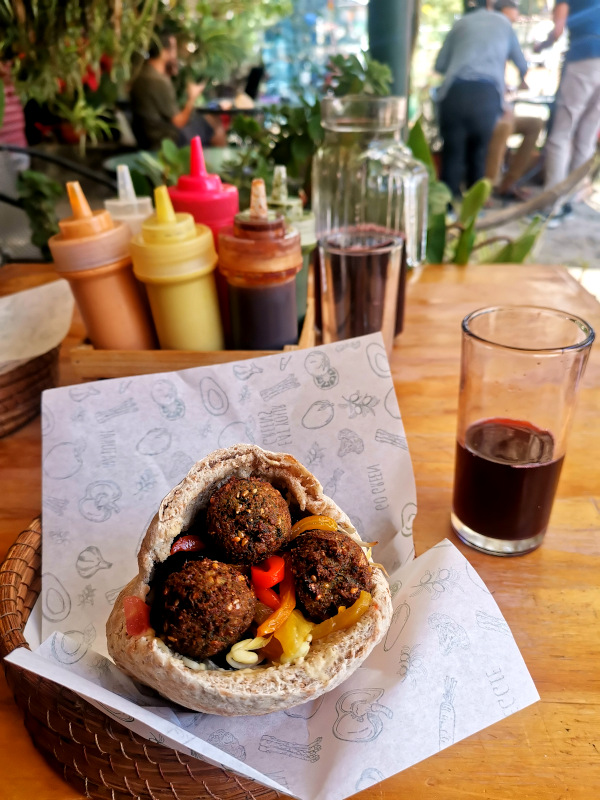 A bowl with a falafel on a table with a basket of sauces behind it and a glass full of red juice
