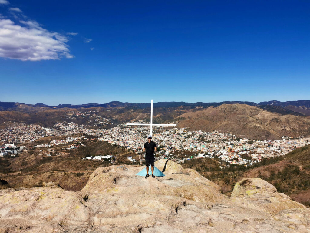 A man in a black t shirt standing in front of a cross at the top of the la bufa guanajuato hike