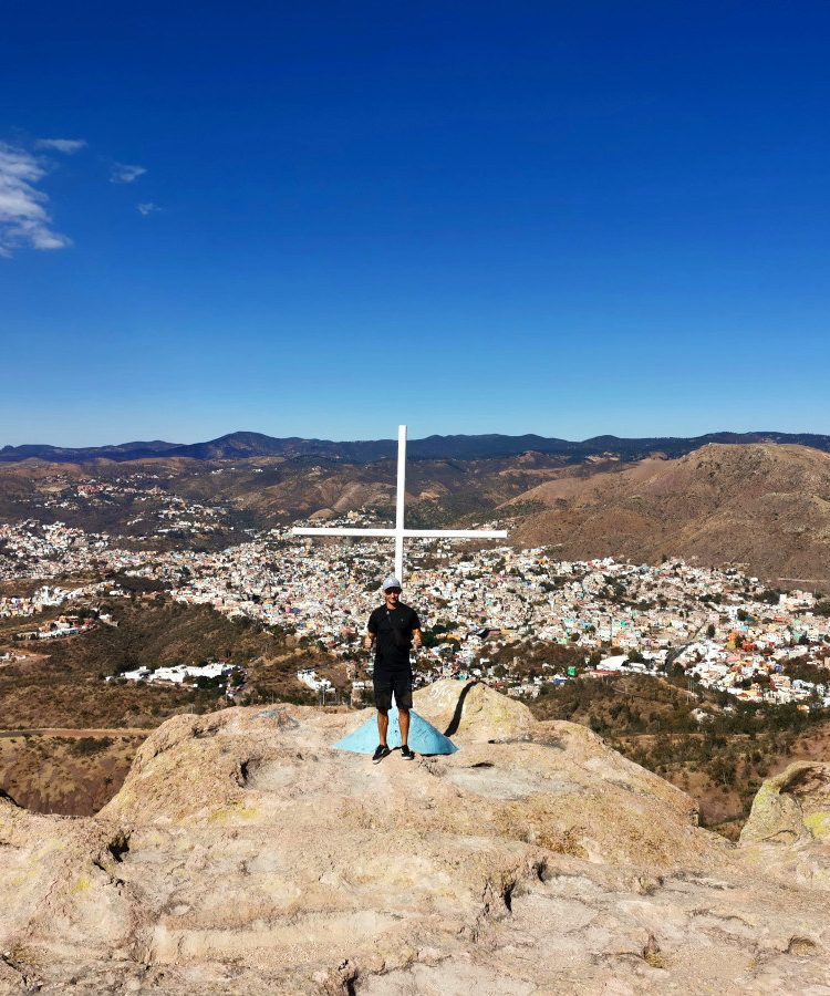 A man in a black t shirt standing in front of a cross at the top of the la bufa guanajuato hike