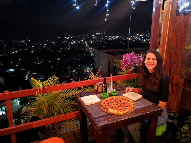 Katharina sitting at a wooden table with a pizza on top and the skyline of Tepoztlán at night in the back 