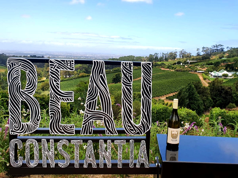 A bottle of wine in front of a sign with vine yards covering the hills behind