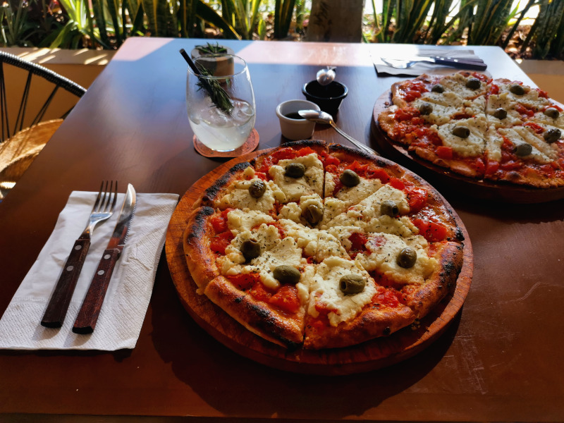 Two pizzas served on a wodden plate with olives and vegan cheese on top at Coral Pizza