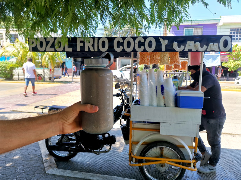 Allan holding a bottle filled with brown Pozol in his hand in front of a blue Pozol stand in Playa del Carmen