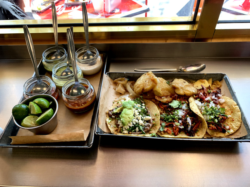Two metal trays covered in glass jars filled with salsa and another tray covered in tacos