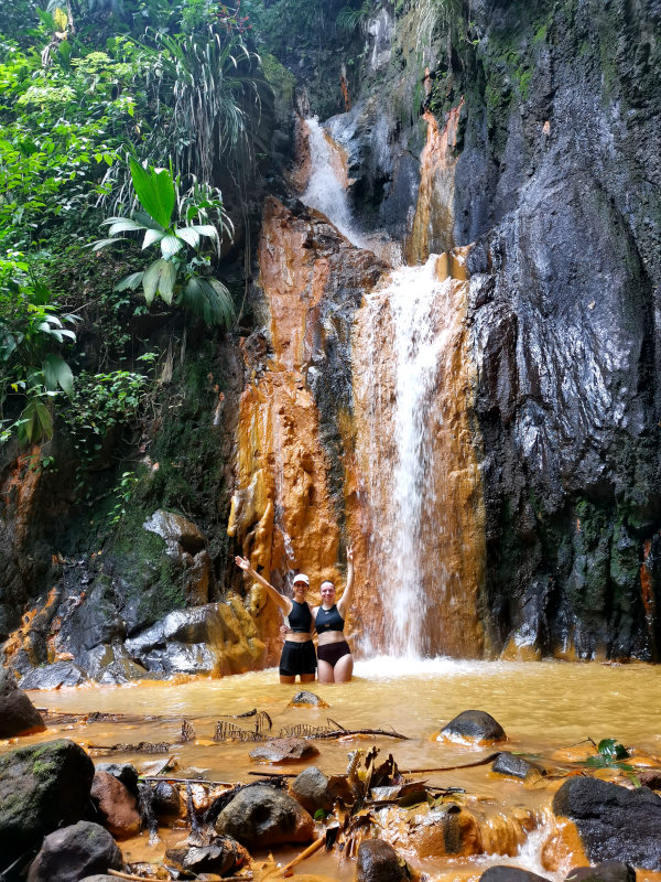 Two woman standing in the water at the bottom of Golden Falls Grenada that is colored brown from the sulphur in the water and one of the best activities in Grenada