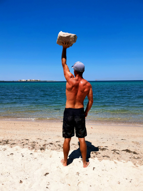 A man in a blue hat and boardshorts working out on the road using a rock at the beach