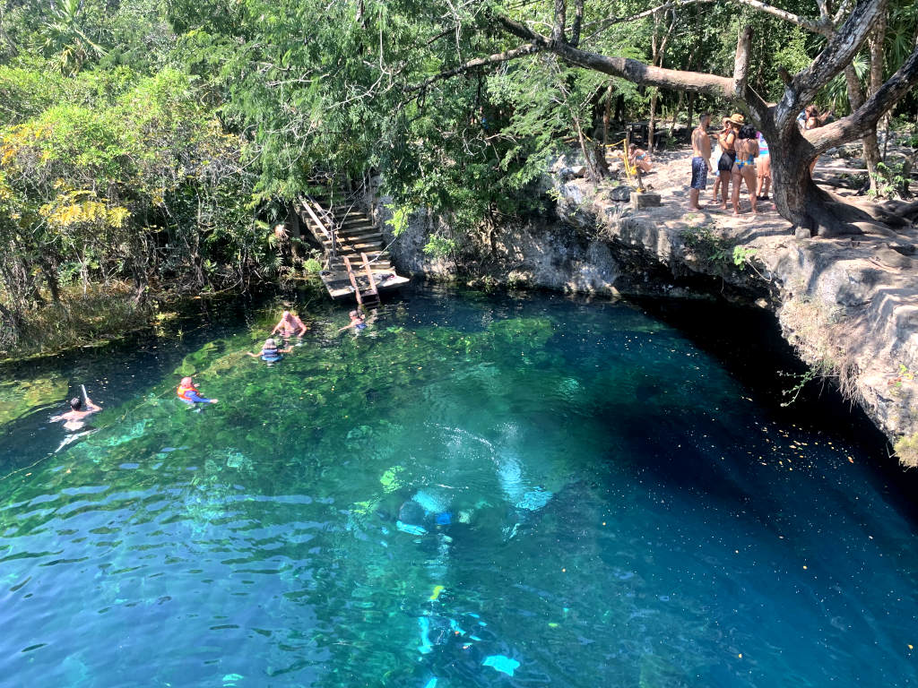 Scuba divers under the water in a cenote with a group of people standing on a rocky ledge above them