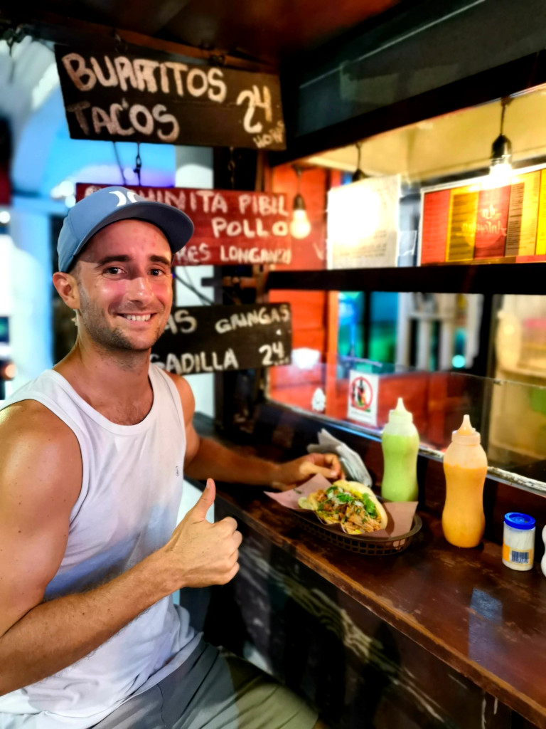 A man in a blue hat and white singlet with his thumb up while eating a cochinita pibil sandwich at la antojera a good food cart for cheap eats in playa del carmen
