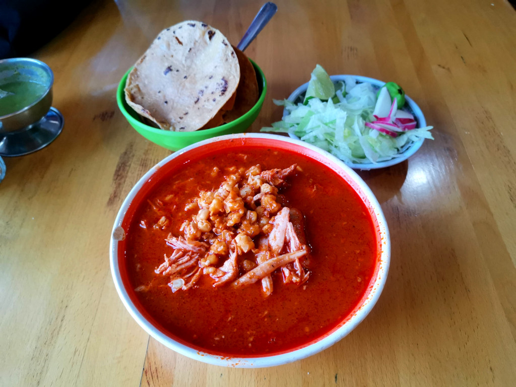 A bowl of bright red Pozole a traditional mexican soup with tostadas and a bowl of salad behind it