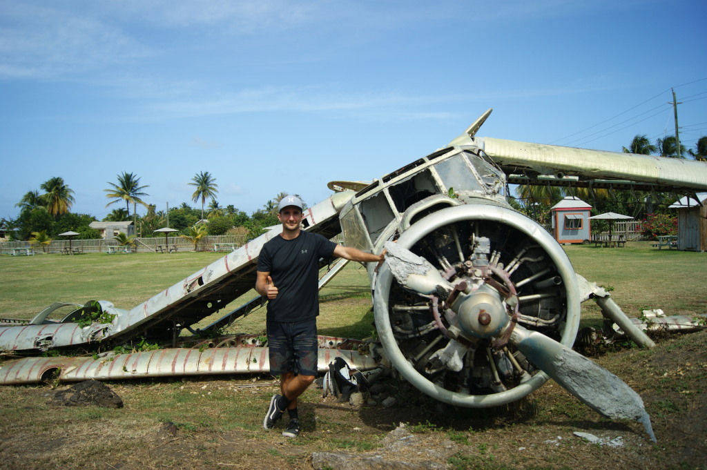 A man in a black t-shirt and shorts standing with his arm resting on the side of an abandoned plane at Pearls Airport a cool thing to do in Grenada