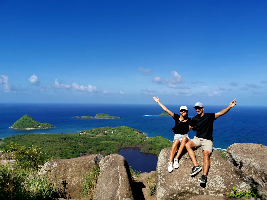 A couple with their arms in the air sitting on a rock with a view of the ocean with some islands behind them