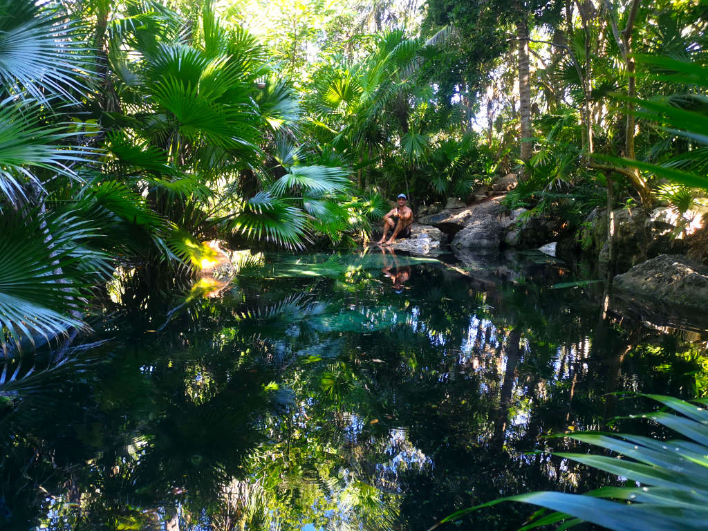 A man in a blue hat sitting next to a cenote in the jungle showing what a cenote is