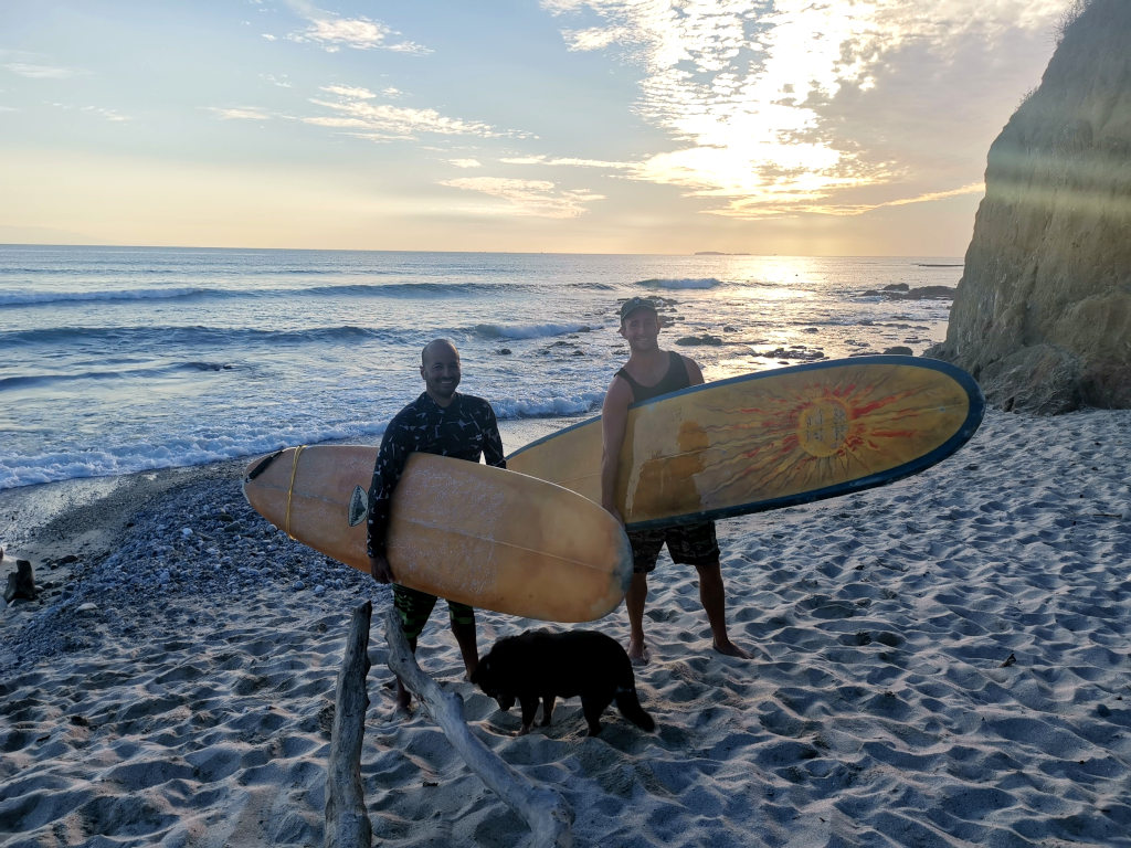 Two men standing on the beach holding surfboards with waves and a sunset behind them