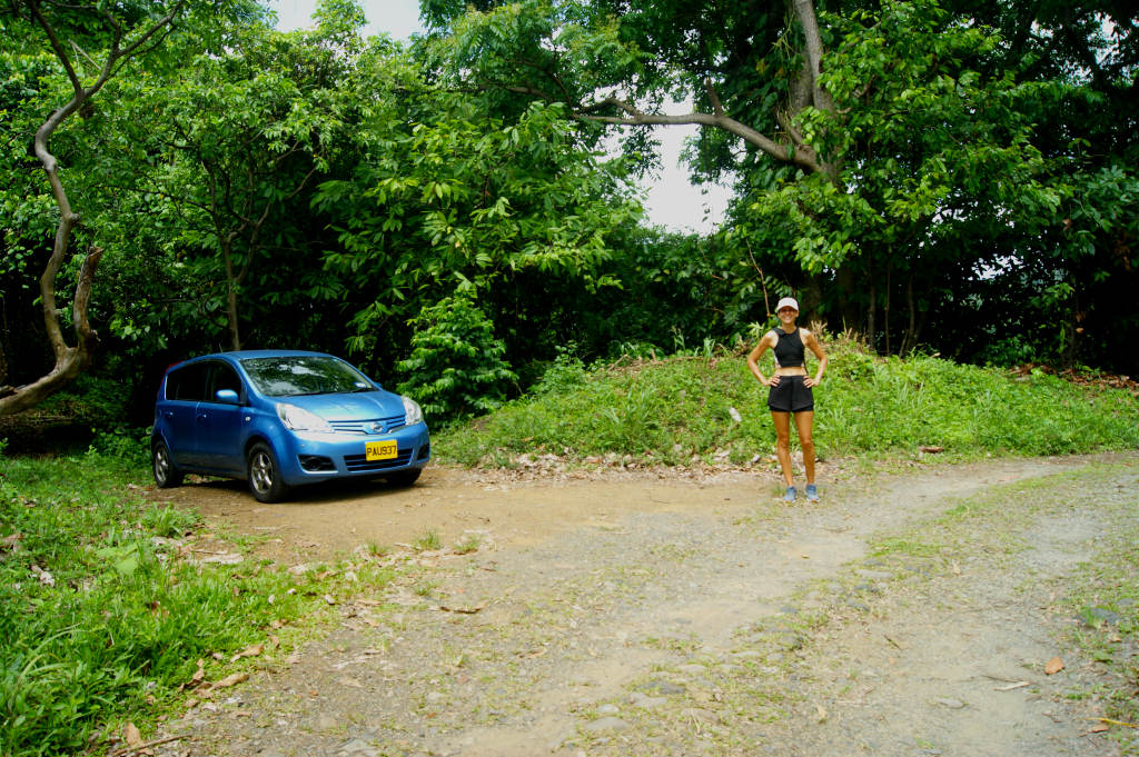 A woman standing on a dirt road next to a blue car at the start of the Mount Saint Catherine Grenada hike starting point