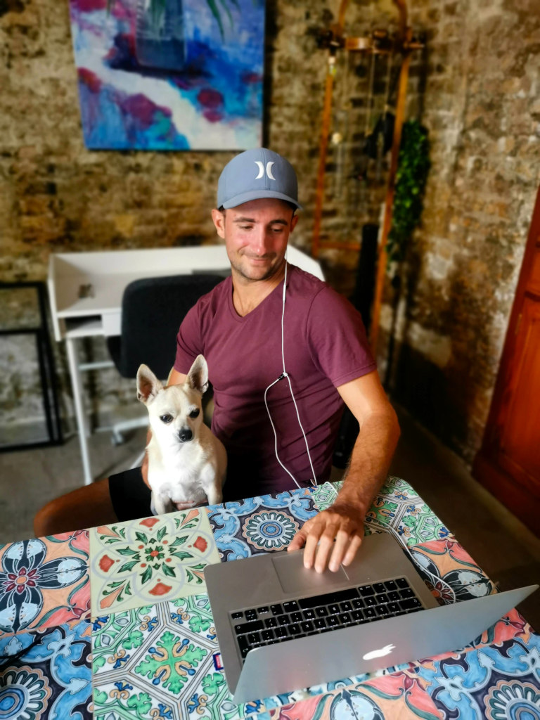 A man in a purple shirt and blue hat working with a chihuahua on his lap while writing a Trusted House Sitters review on his laptop
