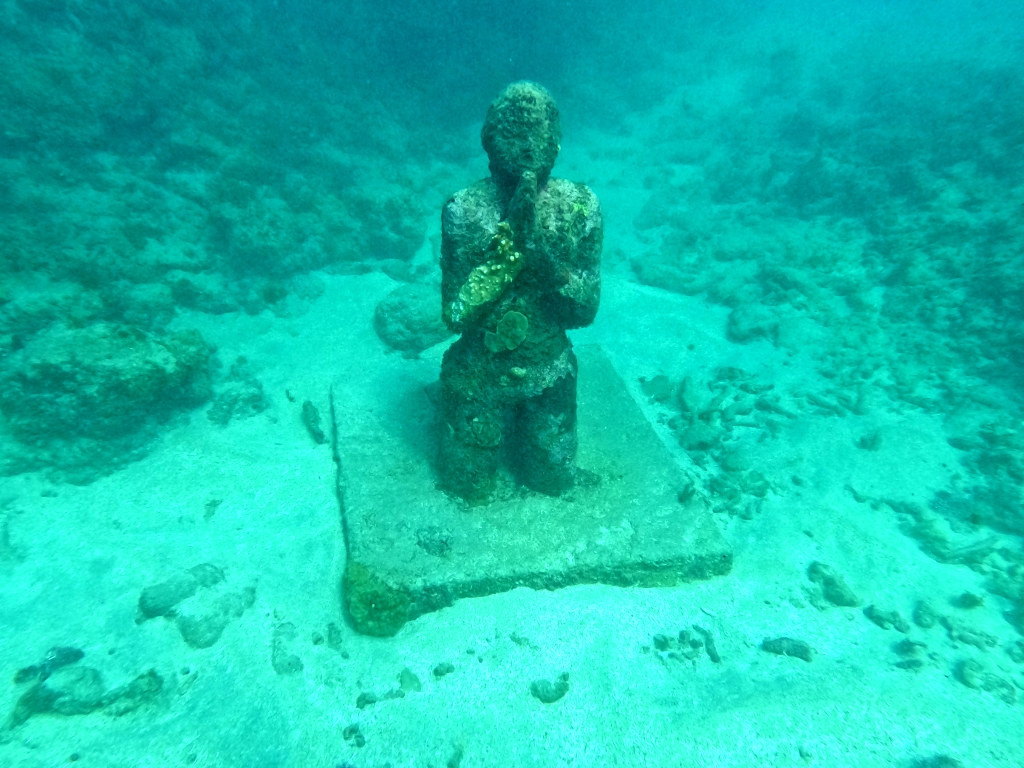 A underwater statue praying on its knees attached to a concrete block on the bottom of the ocean