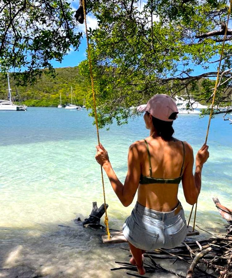 A woman in a bikini top and denim jeans sitting on a swing overlooking clear blue water on hog island grenada