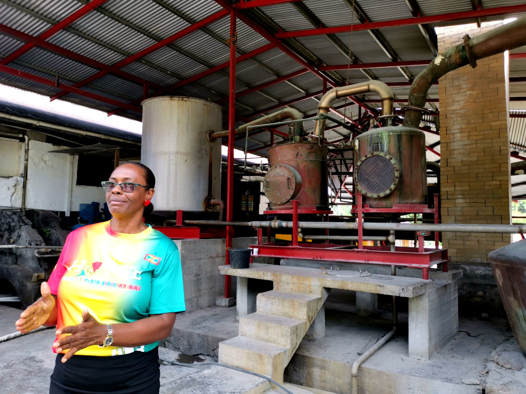 A woman in a red, yellow and green shirt standing in front of machinery at a rum distillery one of the best tours in Grenada
