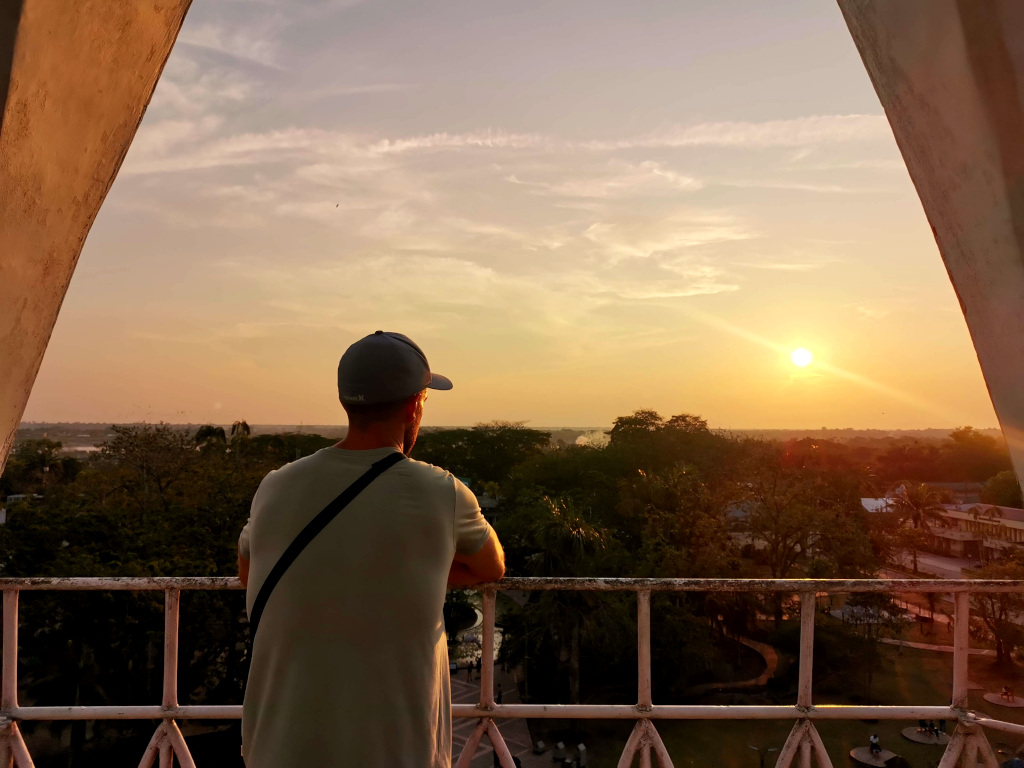 A man in a light green shirt with a blue hat on standing at the top of a church tower watching the sunset in Leticia Colombia