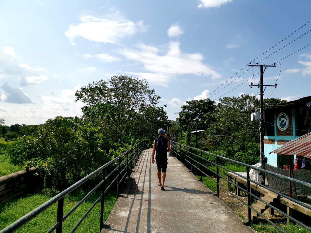 A man in a black singlet with a blue backpack and blue hat on walking along the water in the Colombian Amazon