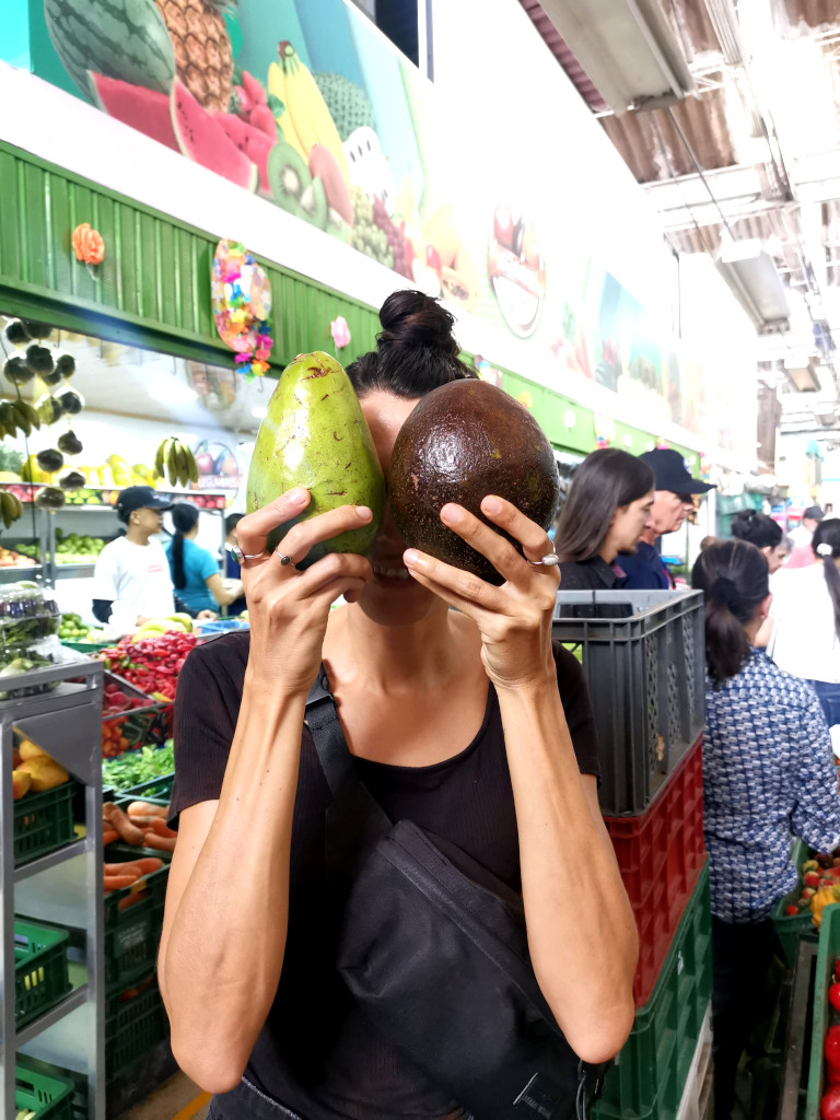 A woman holding two avocados in front of her face at a market