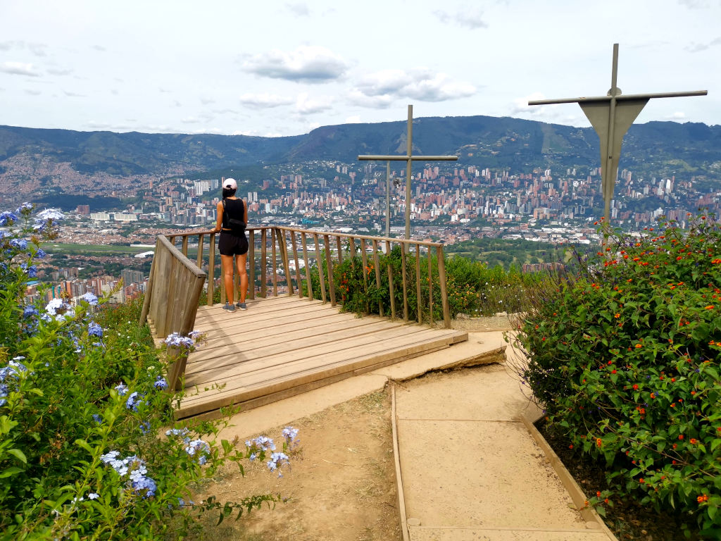 A woman in a pink hat black top and black shorts looking at the view from cerro de las tres cruces one of the best free things to do in Medellin