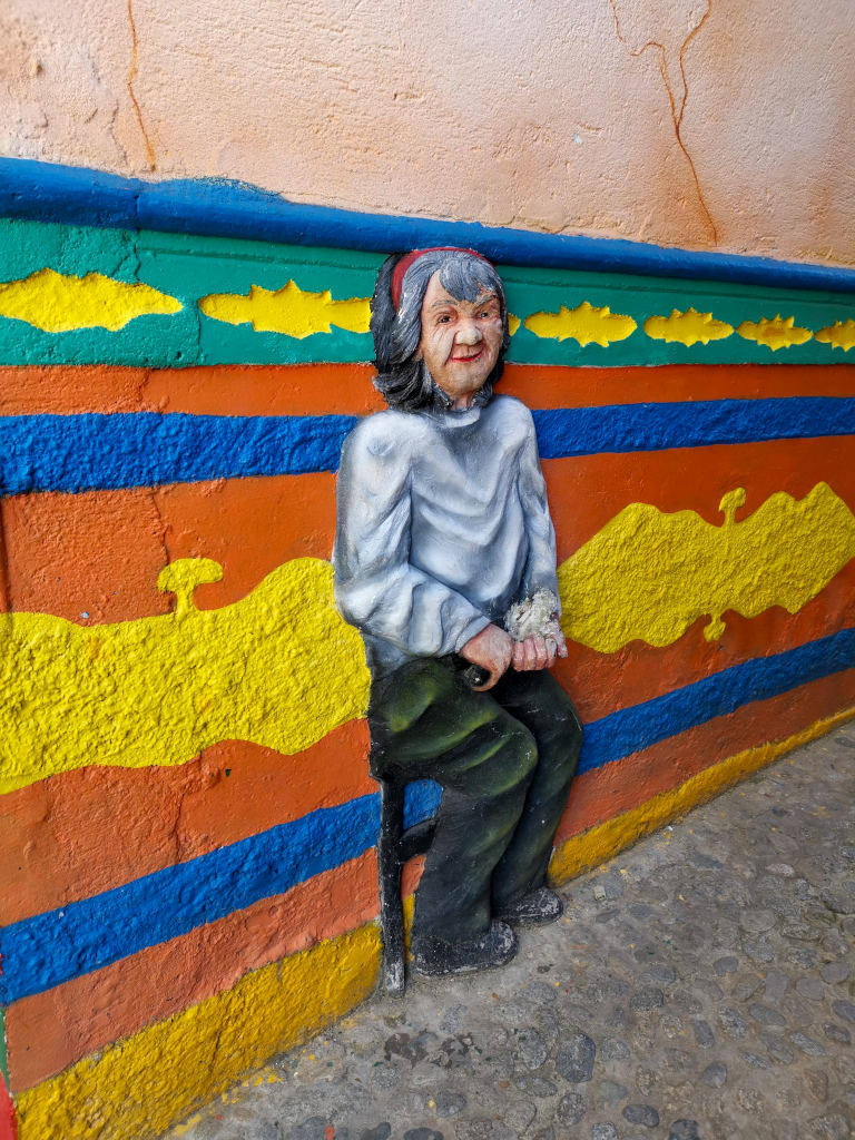 A piece of art on the wall of a building depicting an old woman sitting on a chair