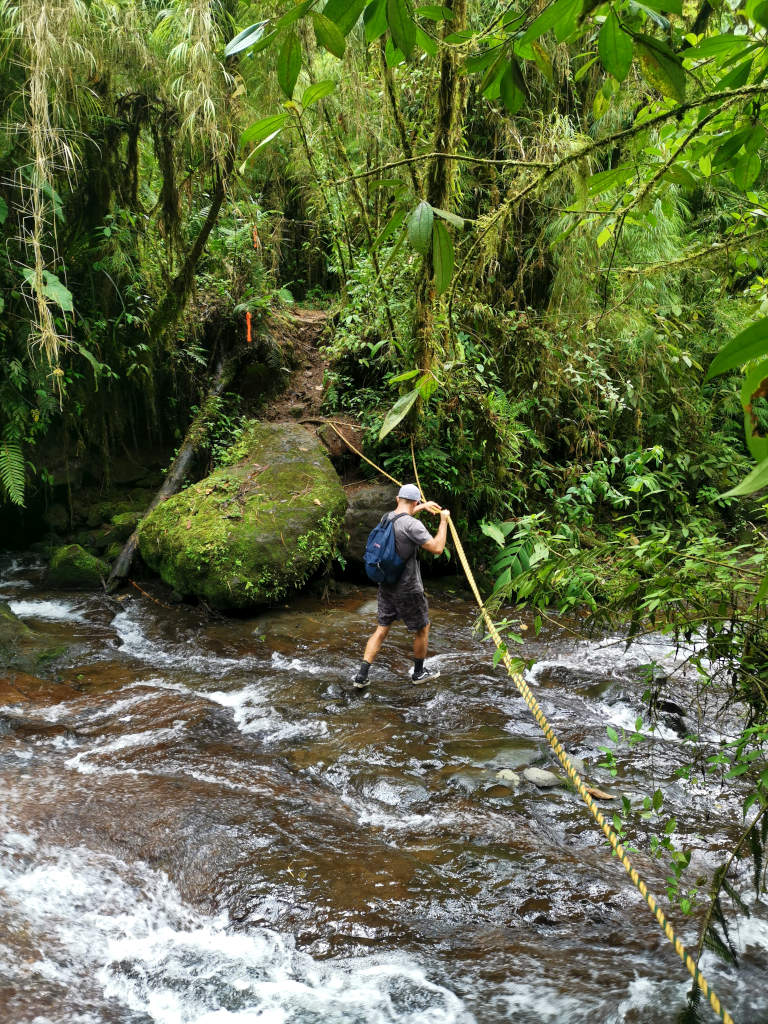 A man in a blue hat with a backpack on using a rope to make his way across a river