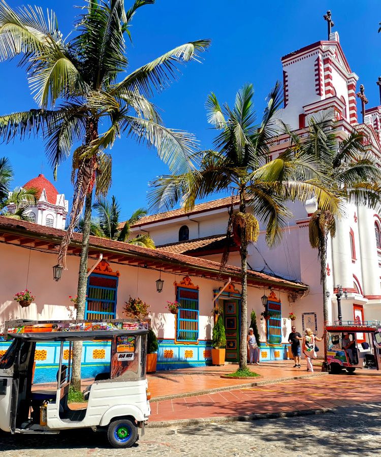 The central square in Guatape Colombia with palm trees and a tuk tuk in front of the red and white church - a must thing to do in Guatape