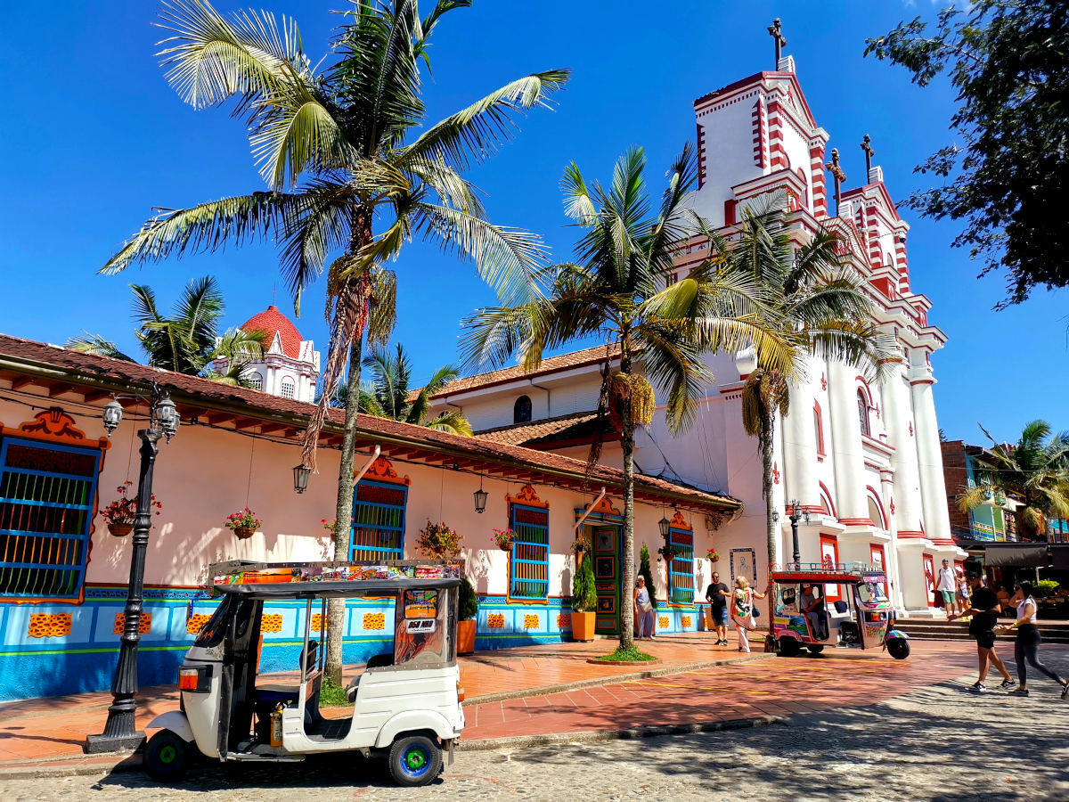 The central square in Guatape Colombia with palm trees and a tuk tuk in front of the red and white church - a must thing to do in Guatape