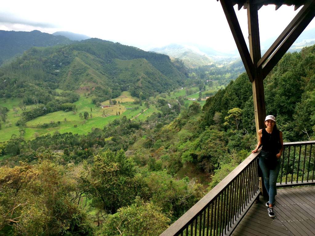 A woman in a pink hat black singlet and jeans standing on a wooden lookout with a view of the cocora valley in the background 