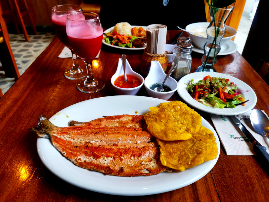 A plate of trout with patacons on a table in a restaurant with salad on a plate and two cups of fruit juice at one of the best restaurants in Jardin Colombia