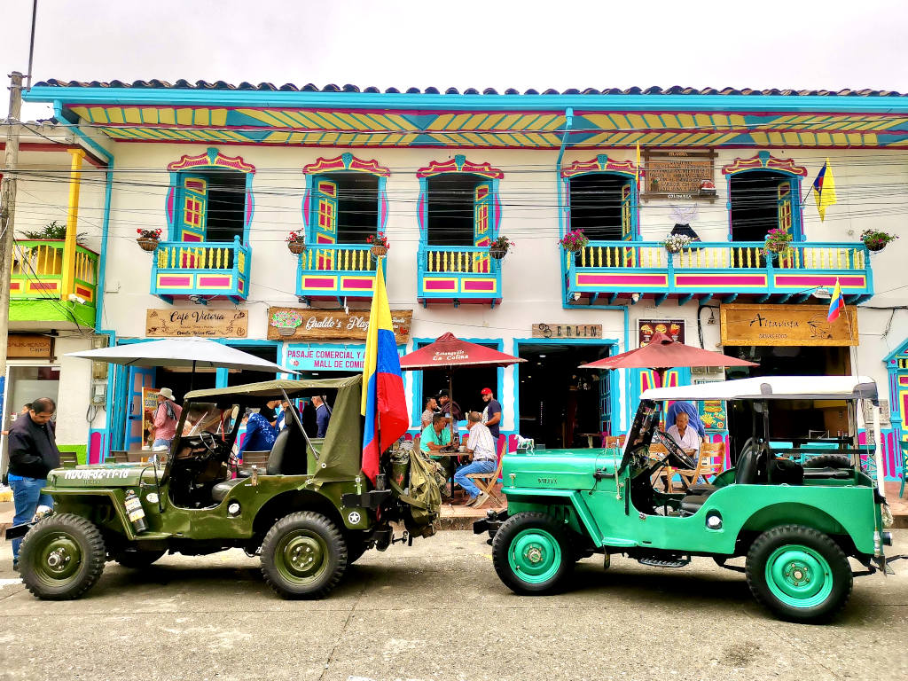 Two colorful jeeps parked on the street at the main square of filandia colombia with colorful buildings int he background and locals sitting on the square on tables at local cafes