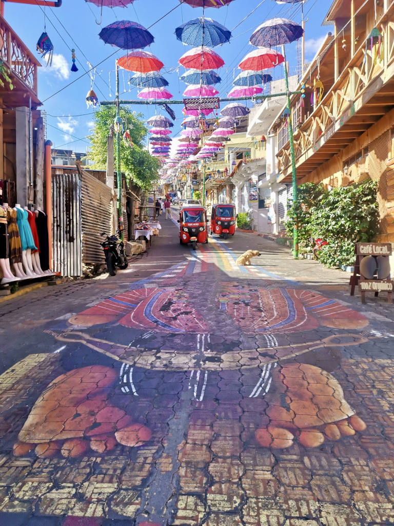 A colorful street with paintings on the ground and colorful umbrellas hanging in the air at the entrance to San Juan La Laguna