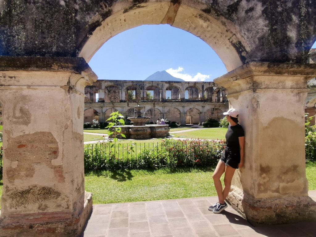 A woman leaning on a wall enjoying the view of some Antigua ruins with a volcano rising up in the air above them