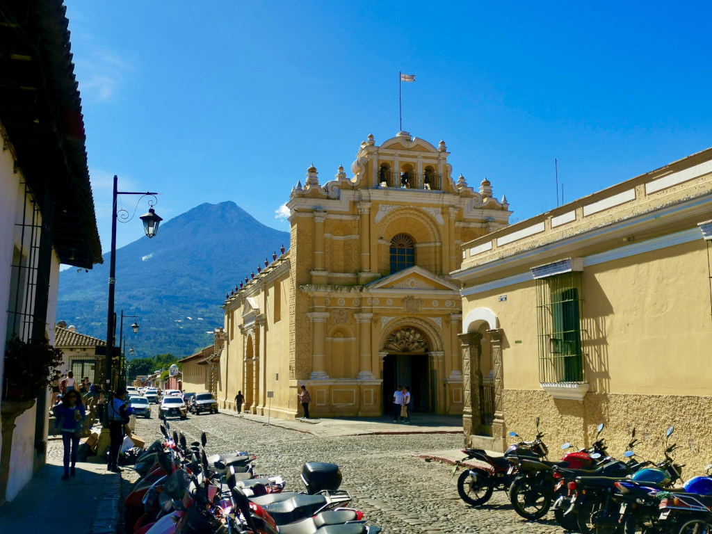 A yellow church on a street in Antigua Guatemala with a volcano rising up behind it