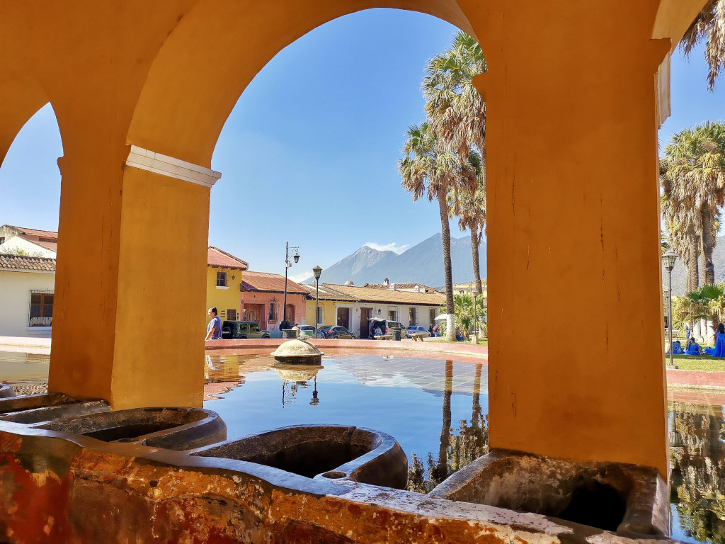 A set of old washing basins in Antigua Guatemala reflecting the background of a volcano, colorful colonial buildings and palm trees