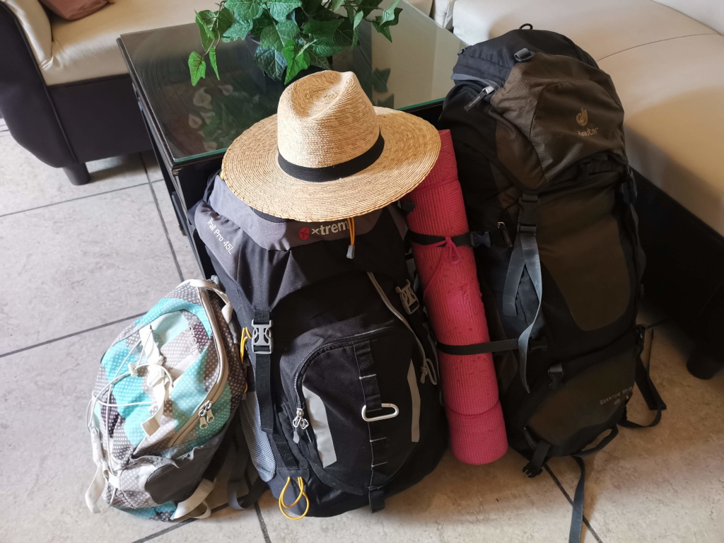 Two big backpacks and a small one with a straw hat on top and a yoga mat attached